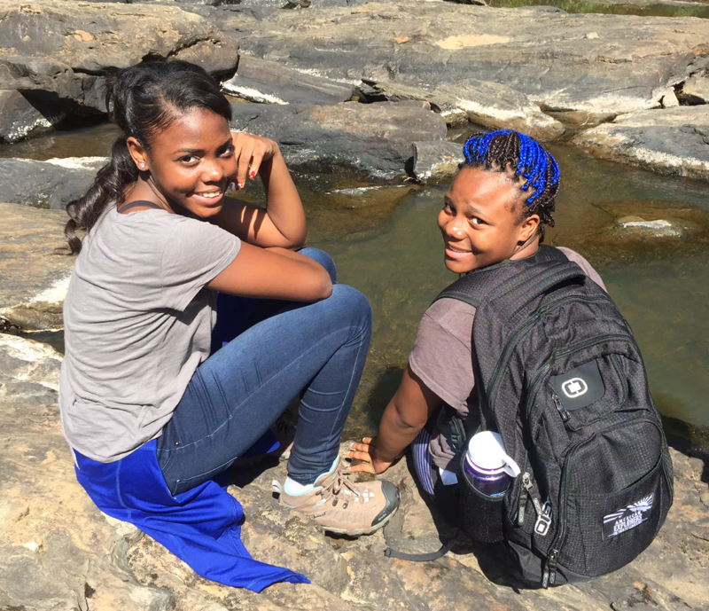 Two young women are sitting on rocks near a stream or river. The woman on the left has dark hair pulled back in a ponytail, a gray t-shirt, jeans, and a blue wrap. She is looking at the camera and smiling. The woman on the right has blue hair, a t-shirt, dark pants, and a black backpack. She is also looking at the camera and smiling. The water is calm and reflects the sky.